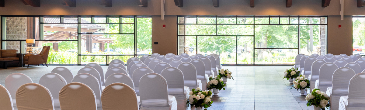 Rows of white-covered chairs line an aisle with floral arrangements, facing large windows.