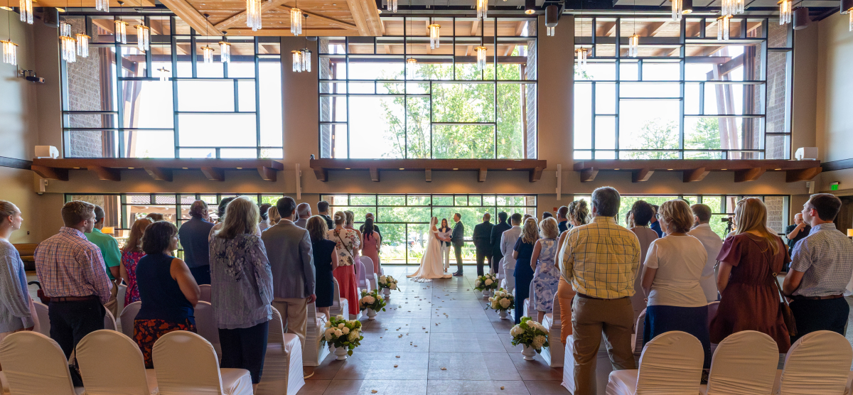 Guests stand at an indoor wedding ceremony, watching the bride and groom at the altar.