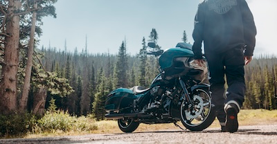 Person walking toward a parked motorcycle on a road with a forest in the background.
