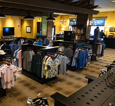 image-card-contact-proshop-396x364 Interior view of a golf pro shop with racks of colorful shirts and golf clubs.
