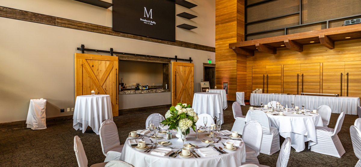 Large event room with many round tables set for a reception, featuring white linens and floral centerpieces.