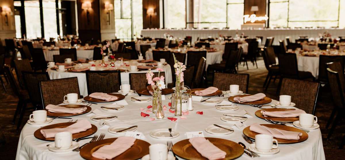 Event room with many round tables set with pink napkins, gold chargers, and small floral centerpieces.