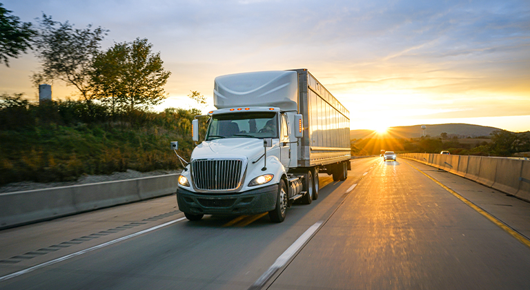 Semi trailer truck driving down highway with sun in background.