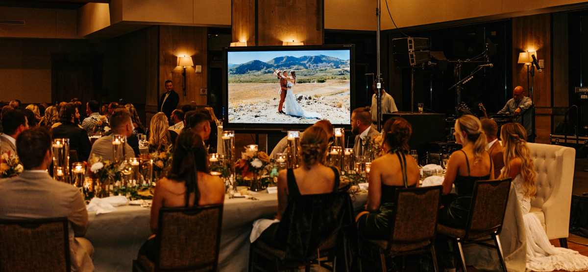 Guests at a reception watch a large screen displaying a wedding photo of a couple in a mountain landscape.