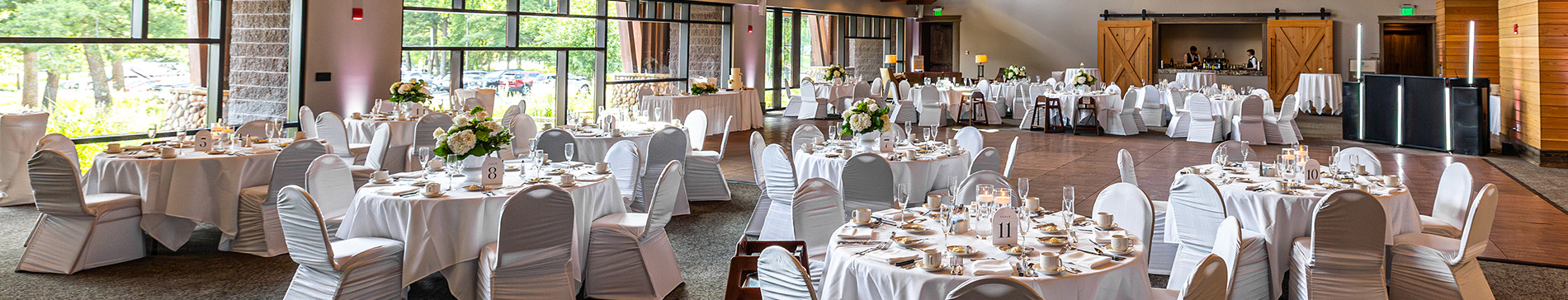 hero-wedding-venue-1900x364 Atrium room with white tables and chairs and a wall of windows behind.