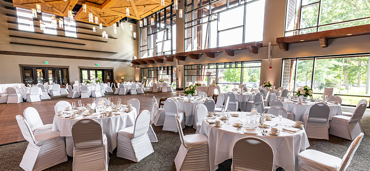carousel-wedding-venue-atrium-1-1200x556 Atrium room with white tables and chairs and a wall of windows behind.