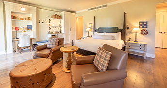 info-card-suites-345x182 Hotel room with one king sized bed and two chairs.