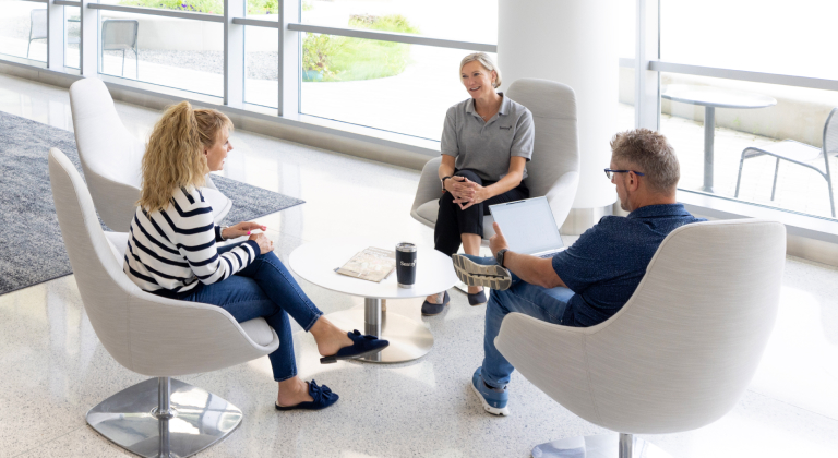 Employees seated and talking in front of a wall of windows. 