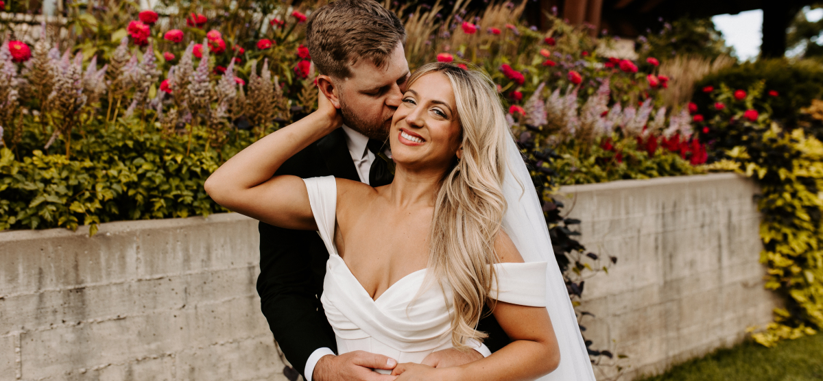 Groom kisses bride’s cheek, bride smiles at camera in front of a flower bed.