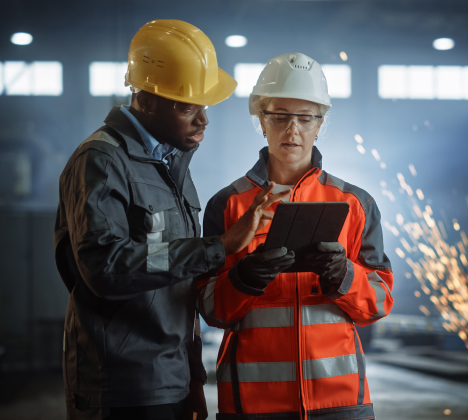 two people working in a metal manufacturing setting wearing hard hats and safety glasses