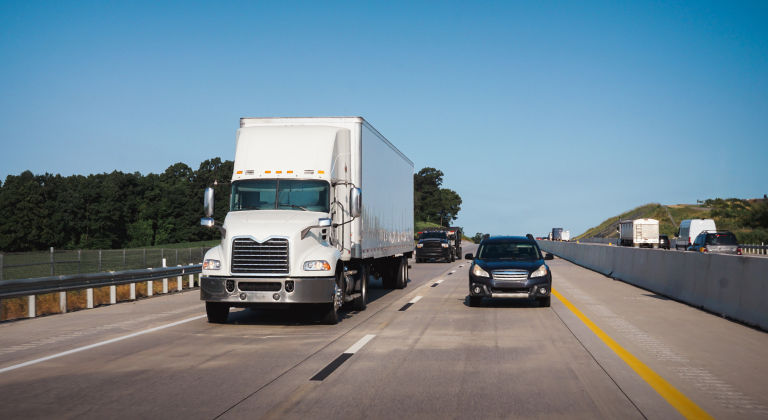 White semi-truck and cars driving on a highway under a clear blue sky.