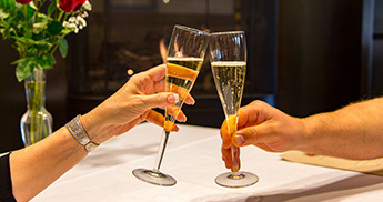 Two people toasting champagne glasses in a restaurant in front of a fire.