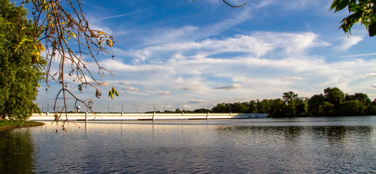 A calm lake with a long white bridge in the distance under a blue sky with clouds.
