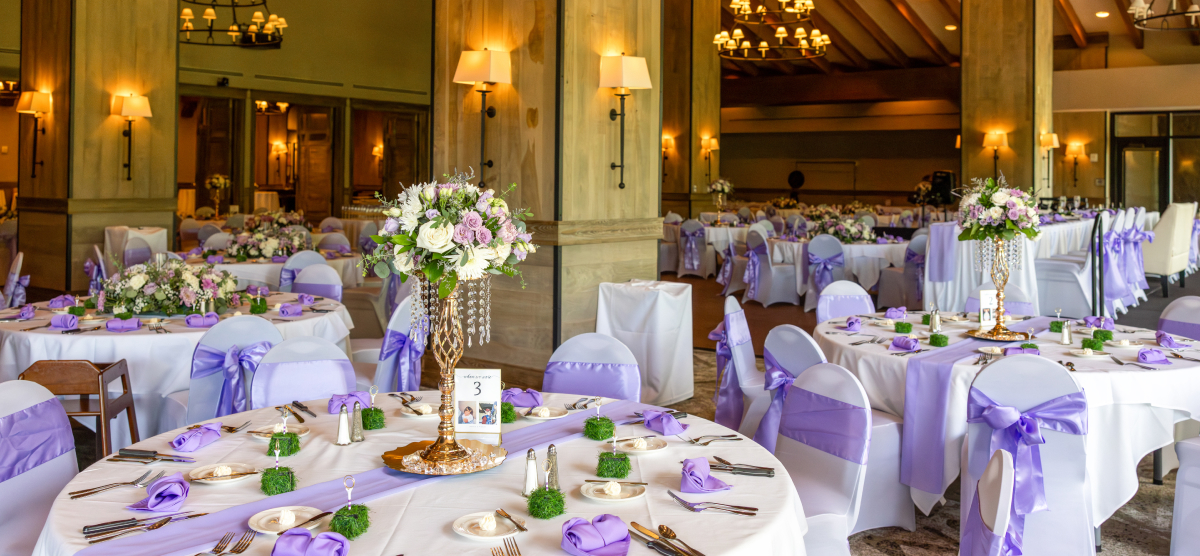 Elegant event room with round tables set with white linens, purple accents, and floral centerpieces.