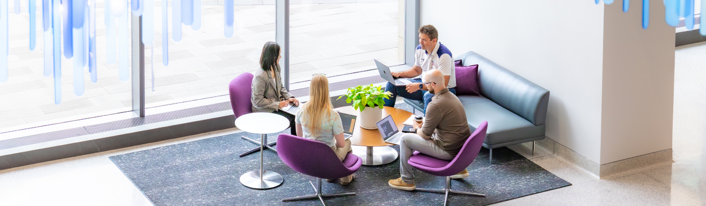 Four colleagues collaborating around a coffee table with laptops in a bright lounge area.