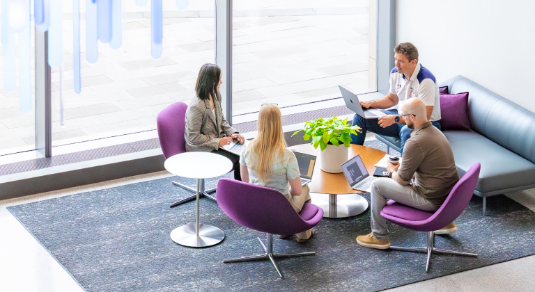 Four colleagues collaborating around a coffee table with laptops in a bright lounge area.
