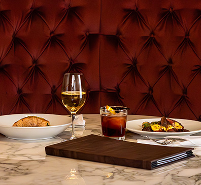 Two plates of food with drinks on a marble table with red fabric bench seat behind. 