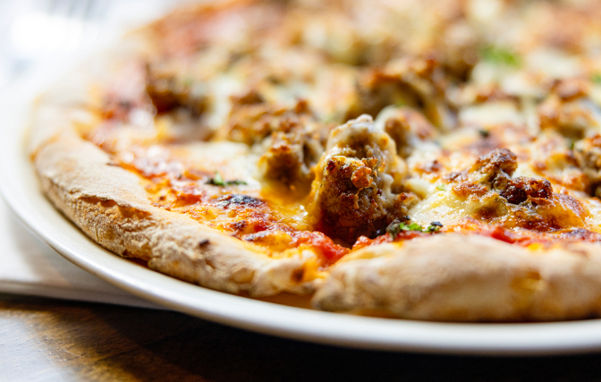 Close-up of a wood fired pizza with melted cheese, tomato sauce, and sausage toppings.