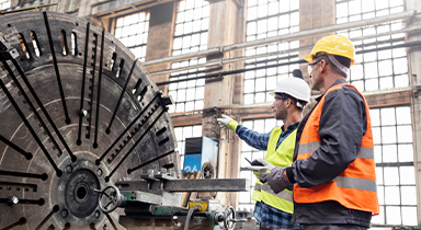 Two men in safety vests reviewing equipment on a manufacturing floor.