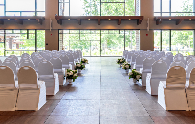 Rows of white-covered chairs line an aisle with floral arrangements, facing large windows.