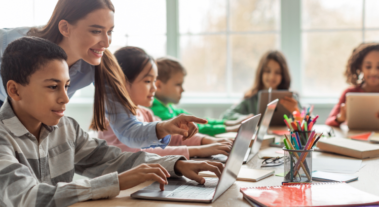 Teacher helps students learn on laptops in a bright classroom.