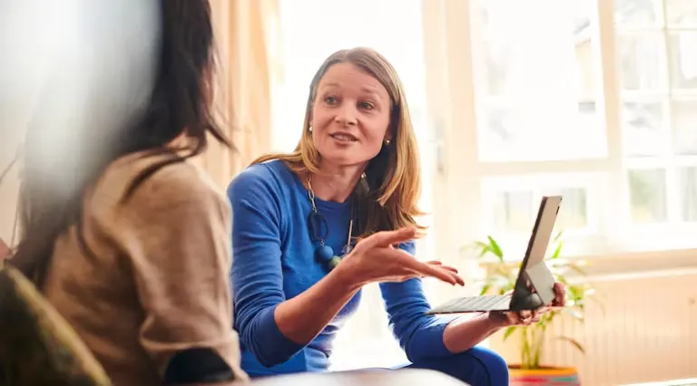 2 women speaking in a living room, one woman is pointing towards an iPad