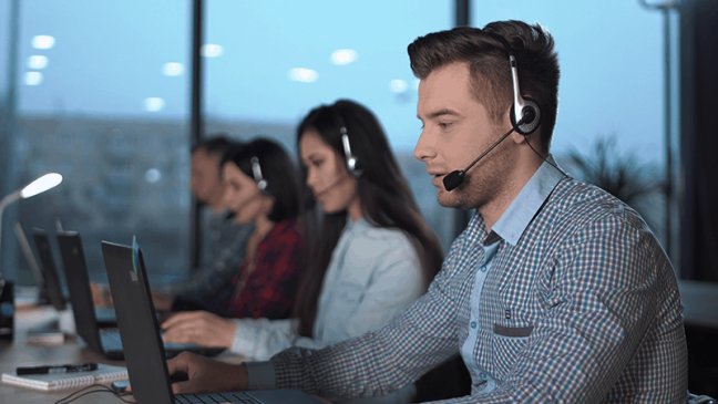 Young men in checked shirt and headset working in call center in modern worldwide office he talking and looking on screen of desktop computer