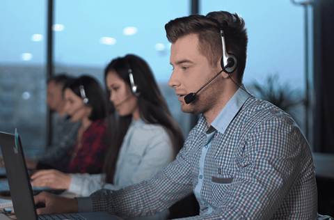 Young men in checked shirt and headset working in call center in modern worldwide office he talking and looking on screen of desktop computer