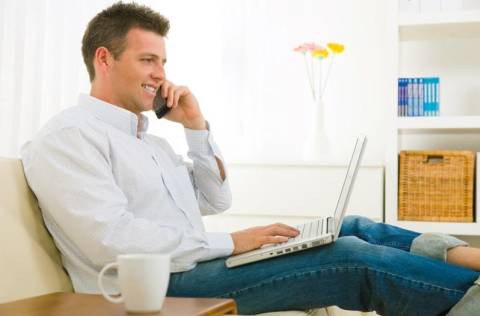 man on couch talking on phone and working on laptop