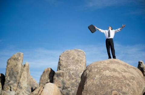 Businessman with briefcase and outstretched arms looking toward sky standing on boulders
