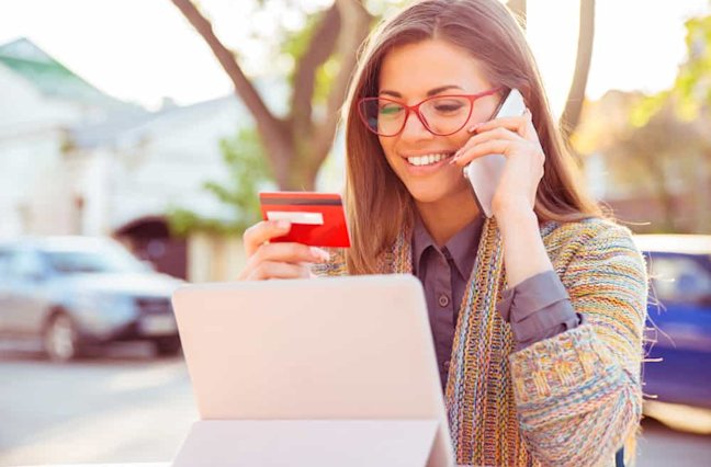 Smiling woman sitting outdoors talking on mobile phone making online payment on her tablet computer outside on a sunny autumn day