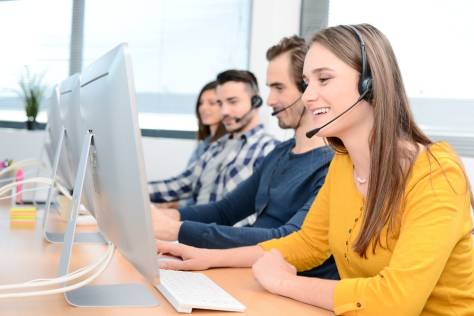portrait of beautiful and cheerful young woman telephone operator with headset working on desktop computer in row in customer service call support helpline business center with co-worker in background