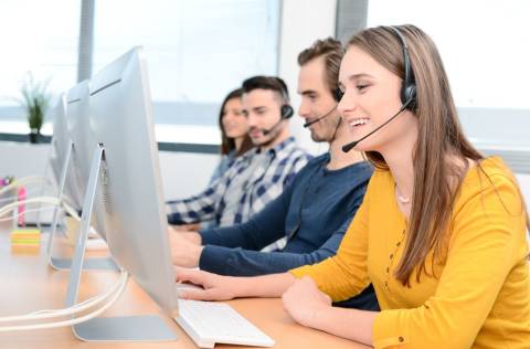 portrait of beautiful and cheerful young woman telephone operator with headset working on desktop computer in row in customer service call support helpline business center with co-worker in background