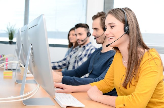 portrait of beautiful and cheerful young woman telephone operator with headset working on desktop computer in row in customer service call support helpline business center with co-worker in background