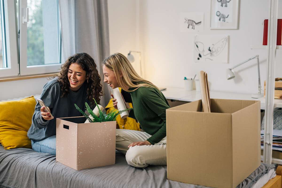 Two girls sit on a bed with moving boxes laughing at a picture