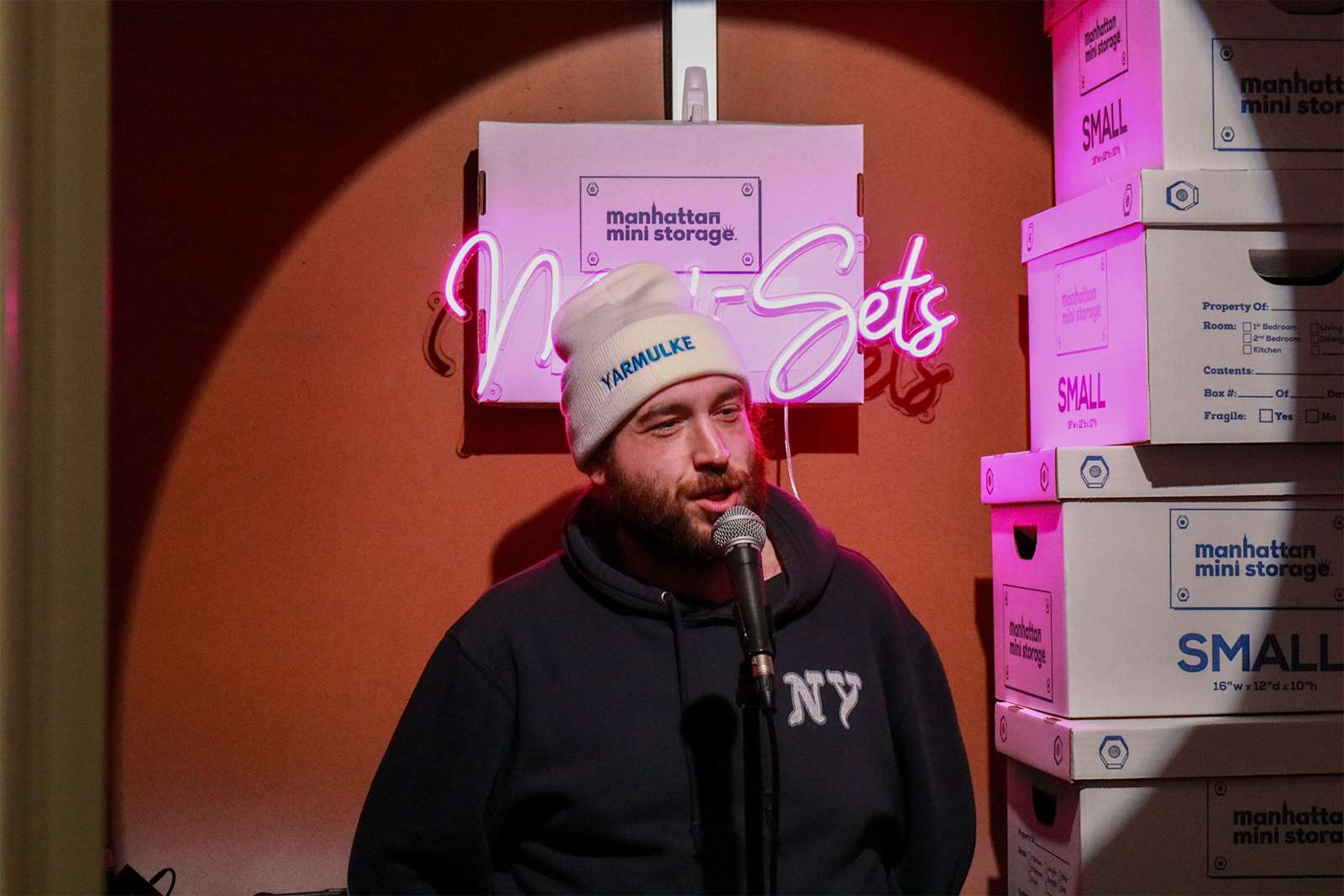 Comedian performing stand-up at a Manhattan Mini Storage event, standing in front of storage boxes with a neon 'Mini Sets' sign in the background.