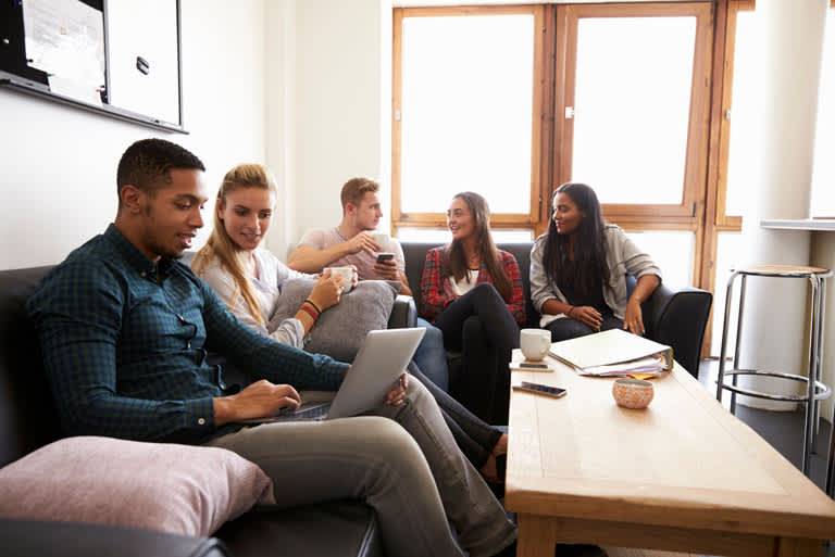 College students gather around a coffee table. Some look at a laptop, while others work on homework