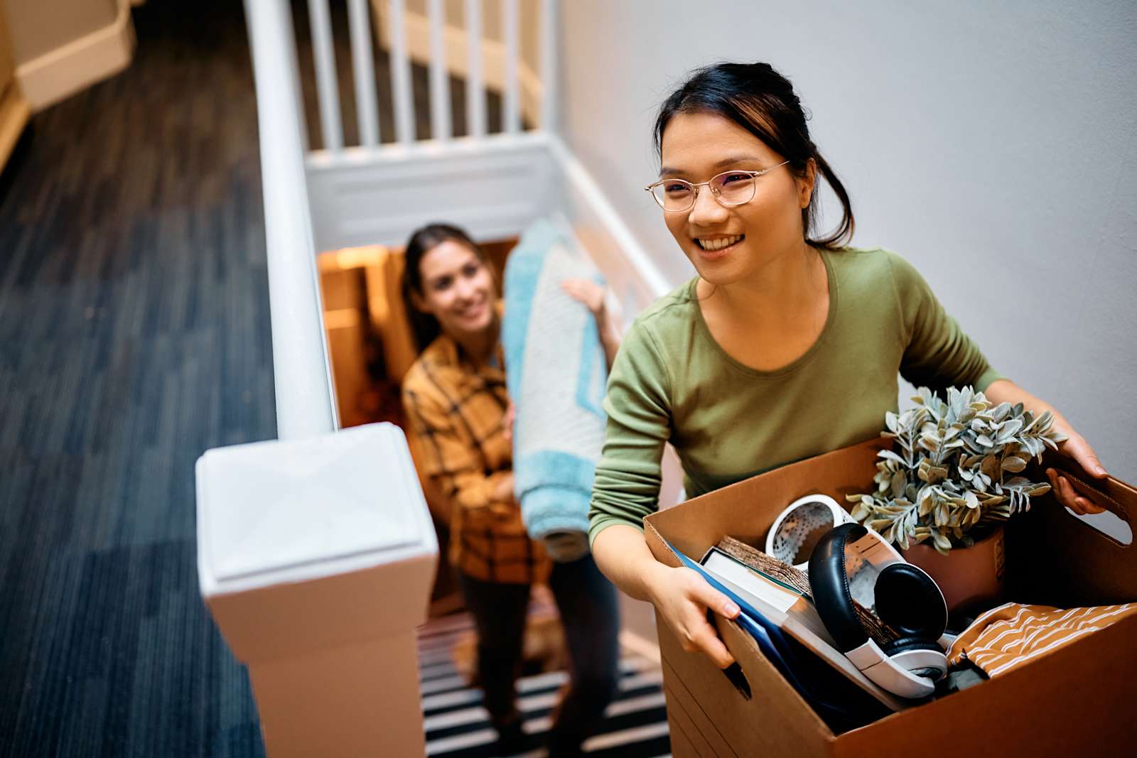 A young woman carries a box of items up a set of stairs while her friend carries a rug behind her.