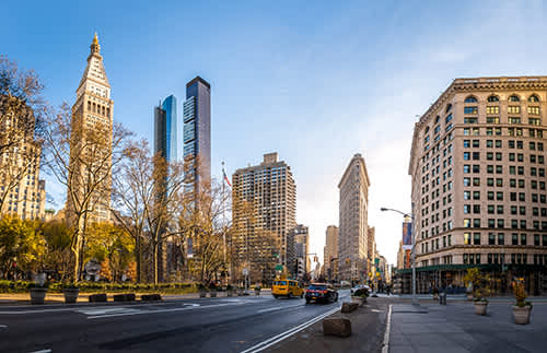 A wide city street lined with tall buildings, including historic and modern skyscrapers, with light traffic and clear skies.