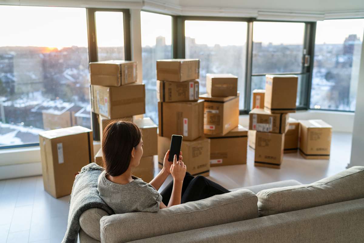 A woman sits on the couch looking at her phone with a stack of moving boxes in front of her