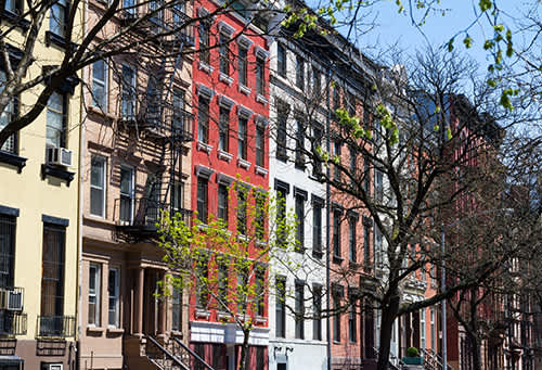 Row of colorful urban brownstones with fire escapes, framed by budding spring trees. The scene conveys a lively, vibrant East Village feel.