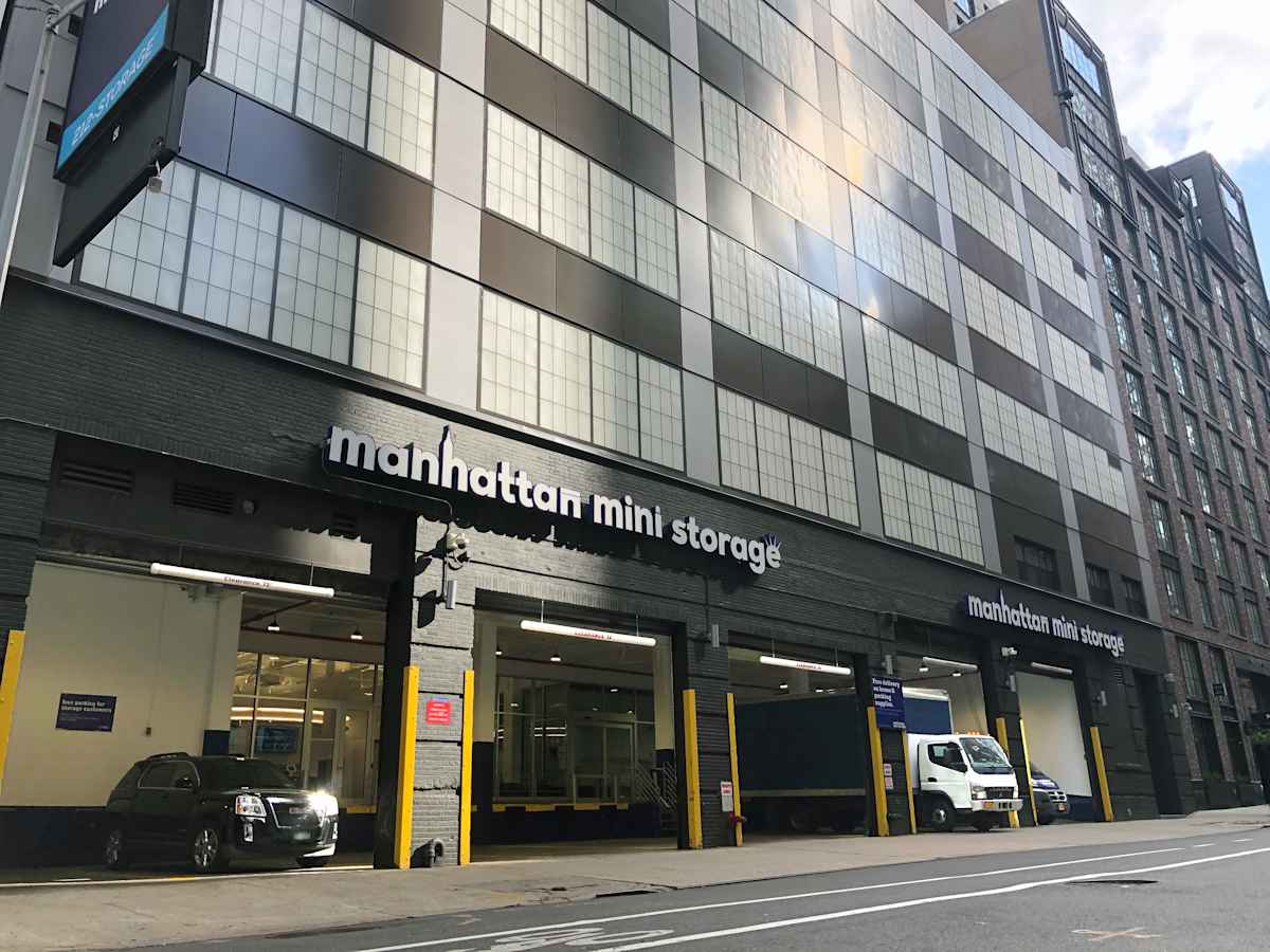 Exterior of Manhattan Mini Storage at 543 West 43rd Street in New York City, showing multiple vehicle bays with open garage doors, a parked black SUV, and a white moving truck under a modern industrial-style building.