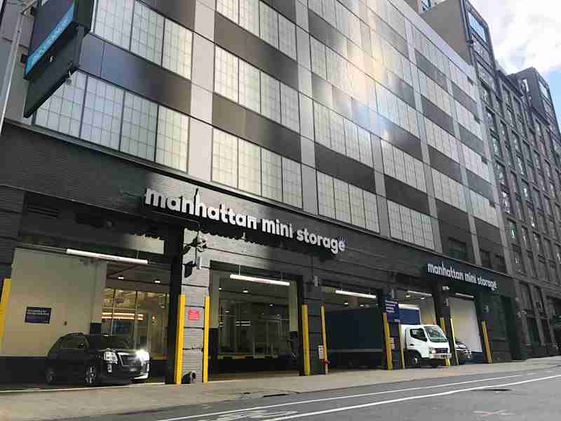 Exterior of Manhattan Mini Storage at 543 West 43rd Street in New York City, showing multiple vehicle bays with open garage doors, a parked black SUV, and a white moving truck under a modern industrial-style building.