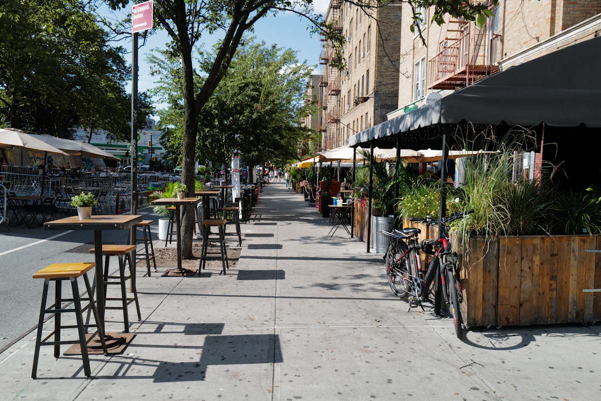 Sidewalk with outdoor dining, tall stools, trees, and bikes on Dyckman Street, Inwood NYC.