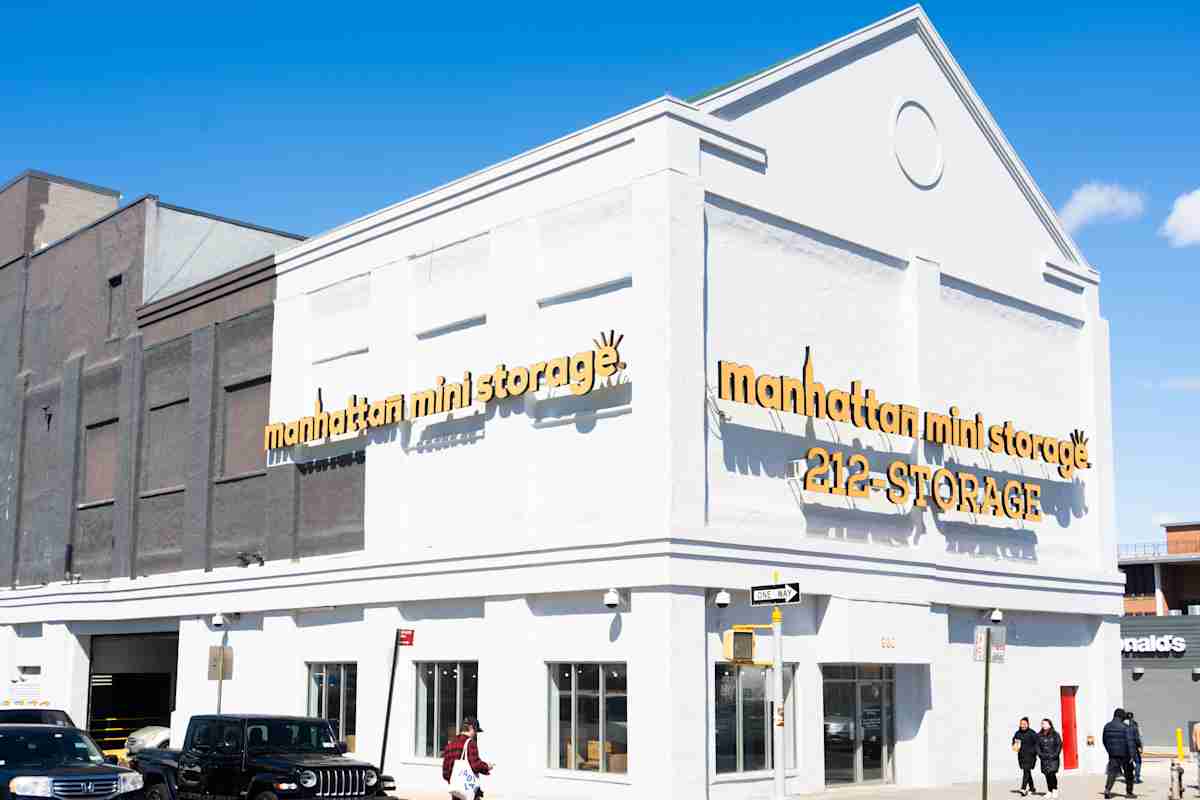 Corner view of a Manhattan Mini Storage facility on 4th and 38th with bold yellow signage and phone number 212-STORAGE on a bright white building, located on a city street with pedestrians and parked vehicles.