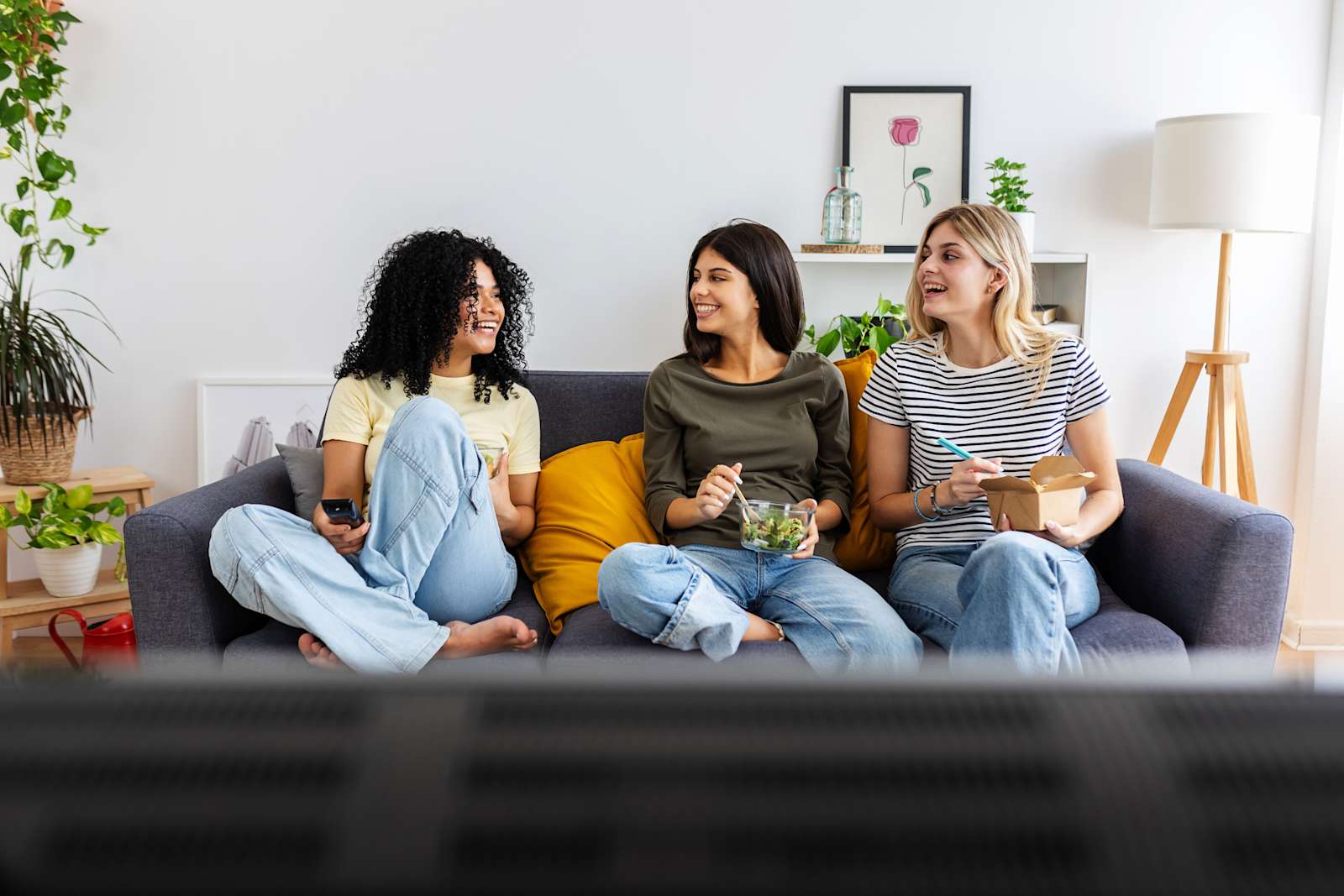 Three young women sit on a couch eating food and laughing