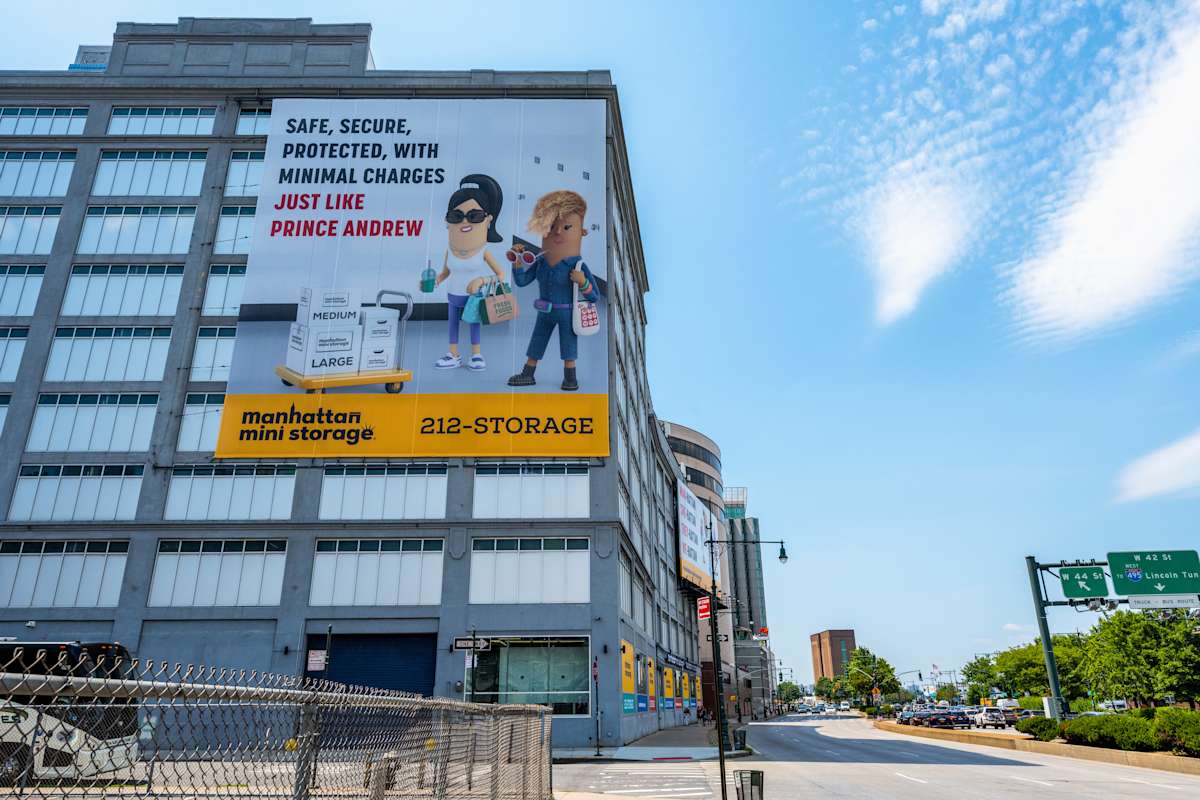 A large advertisement is displayed on the side of a multi-story building for Manhattan Mini Storage. The billboard features cartoon-style figures and reads: SAFE, SECURE, PROTECTED, WITH MINIMAL CHARGES JUST LIKE PRINCE ANDREW.