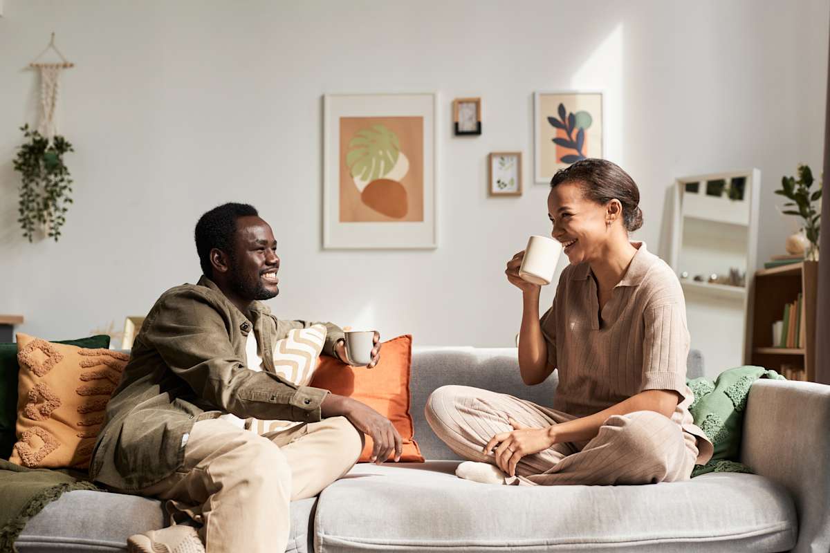 A man and a woman sit on a couch drinking coffee laughing together