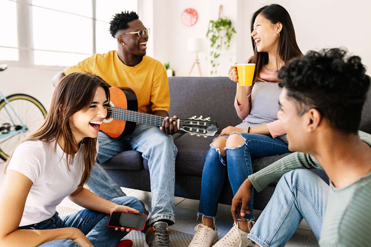 Four young people sit around a couch. one man plays a guitar, one woman drinks a coffee, and all are laughing.