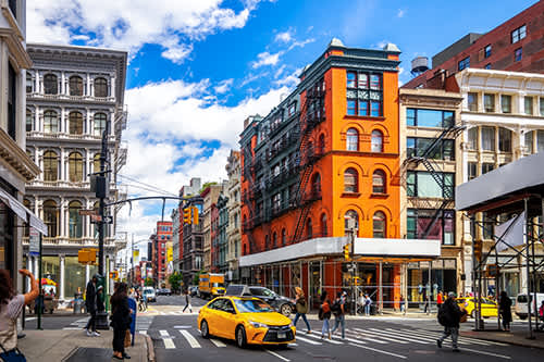 Soho street scene with a vibrant orange building, surrounded by historic architecture. Pedestrians and a yellow taxi create a lively, bustling atmosphere.